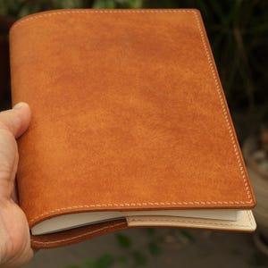 Può includere: Una copertina rettangolare per taccuino realizzata in pelle color cuoio con una superficie testurizzata. La copertina presenta cuciture visibili di colore chiaro lungo i bordi e una sezione in pelle più chiara, color crema, sul bordo inferiore.