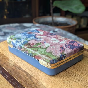 May include: A rectangular blue box with a floral patterned lid. The lid features a mix of pink, blue, and green floral designs. The box has gold-colored trim and is set on a wooden surface.