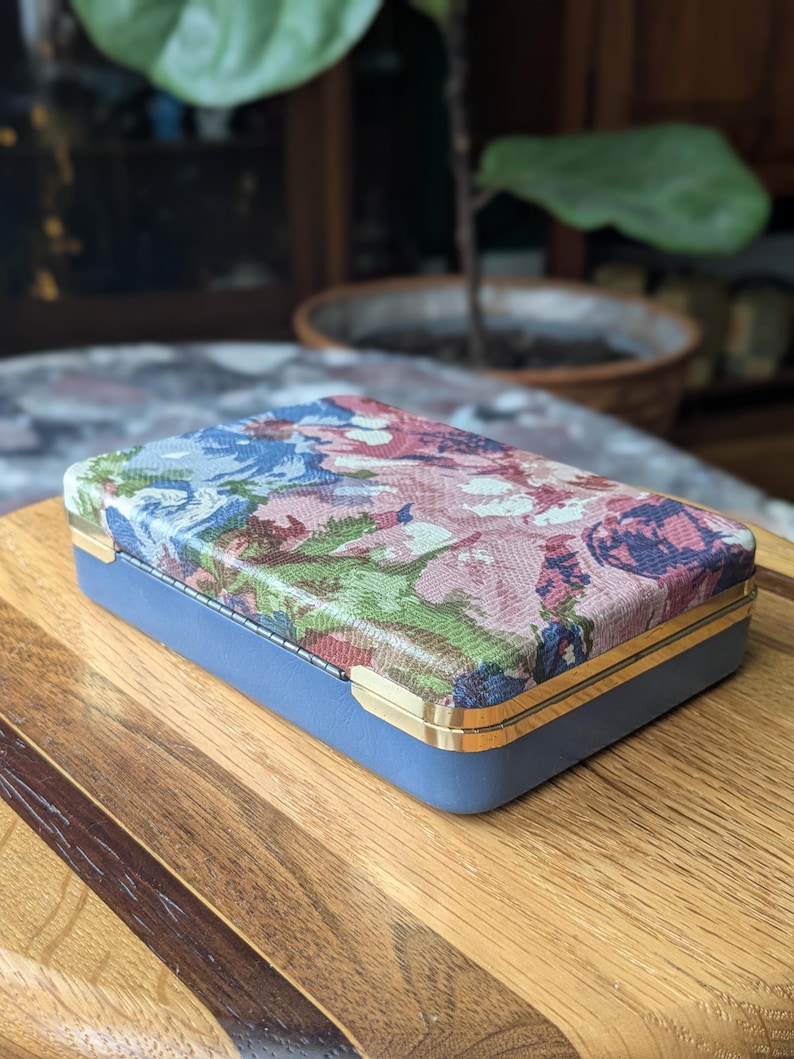 May include: A rectangular blue box with a floral patterned lid. The lid features a mix of pink, blue, and green floral designs. The box has gold-colored trim and is set on a wooden surface.