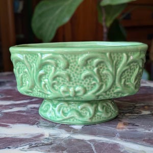 May include: A light green ceramic planter with a pedestal base. The planter features an embossed floral design and a rectangular shape. The planter is sitting on a marble surface.