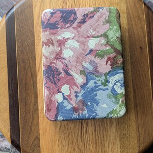 May include: Rectangular floral patterned compact mirror with a gold trim. The mirror features a design of pink, blue, and green flowers. The mirror is resting on a wooden cutting board.