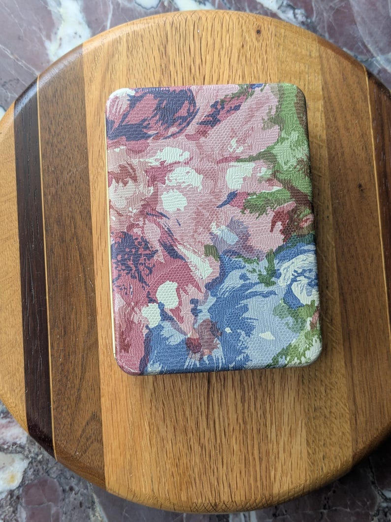May include: Rectangular floral patterned compact mirror with a gold trim. The mirror features a design of pink, blue, and green flowers. The mirror is resting on a wooden cutting board.