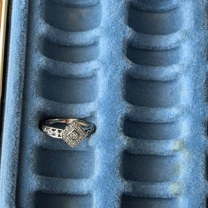 May include: A silver ring with a square, diamond-shaped center stone and intricate detailing. The ring is displayed in a blue velvet jewelry box with multiple ring slots, showcasing its design and craftsmanship.