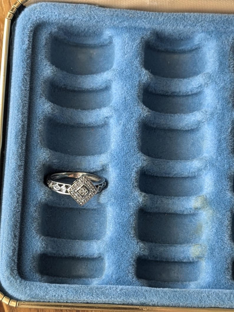 May include: A silver ring with a square, diamond-shaped center stone and intricate detailing. The ring is displayed in a blue velvet jewelry box with multiple ring slots, showcasing its design and craftsmanship.