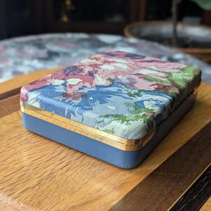 May include: A rectangular box with a floral patterned lid in shades of pink, blue, and green. The box has a blue base with gold trim. The box is sitting on a wooden surface.
