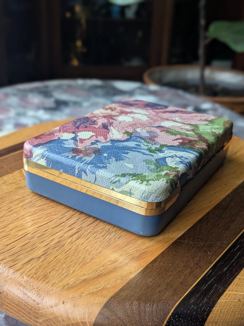 May include: A rectangular box with a floral patterned lid in shades of pink, blue, and green. The box has a blue base with gold trim. The box is sitting on a wooden surface.