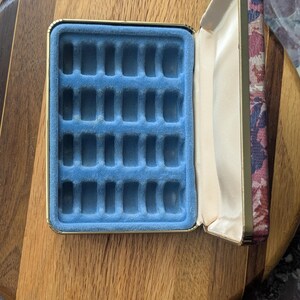 May include: Open jewelry box with a blue velvet interior and a floral patterned lid. The box is rectangular with a gold trim and is set on a wooden surface. The interior has multiple slots for storing jewelry.