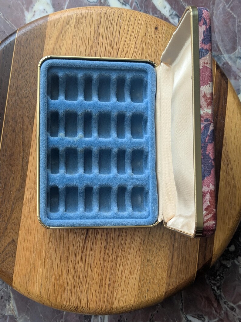 May include: Open jewelry box with a blue velvet interior and a floral patterned lid. The box is rectangular with a gold trim and is set on a wooden surface. The interior has multiple slots for storing jewelry.