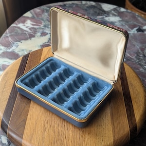 May include: Open blue jewelry box with a gold trim and a floral patterned lid. The interior features a light blue velvet lining with multiple compartments for rings and other small items. The box is placed on a wooden surface.