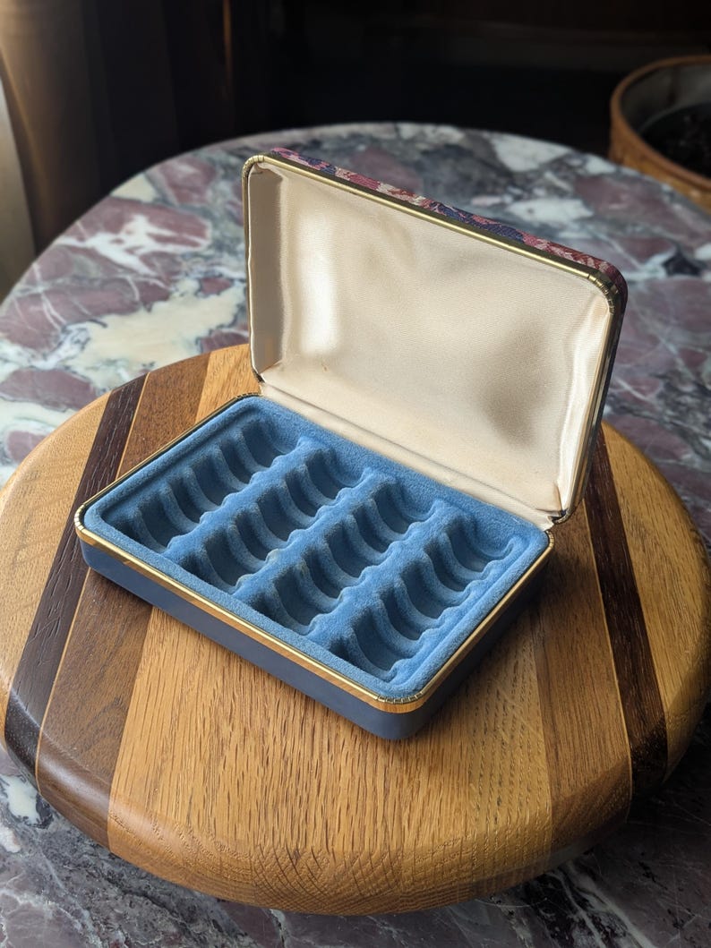 May include: Open blue jewelry box with a gold trim and a floral patterned lid. The interior features a light blue velvet lining with multiple compartments for rings and other small items. The box is placed on a wooden surface.