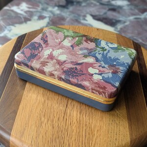 May include: A rectangular box with a floral pattern lid in shades of pink, blue, and green. The box has a navy blue base with gold trim. The box is resting on a wooden cutting board.