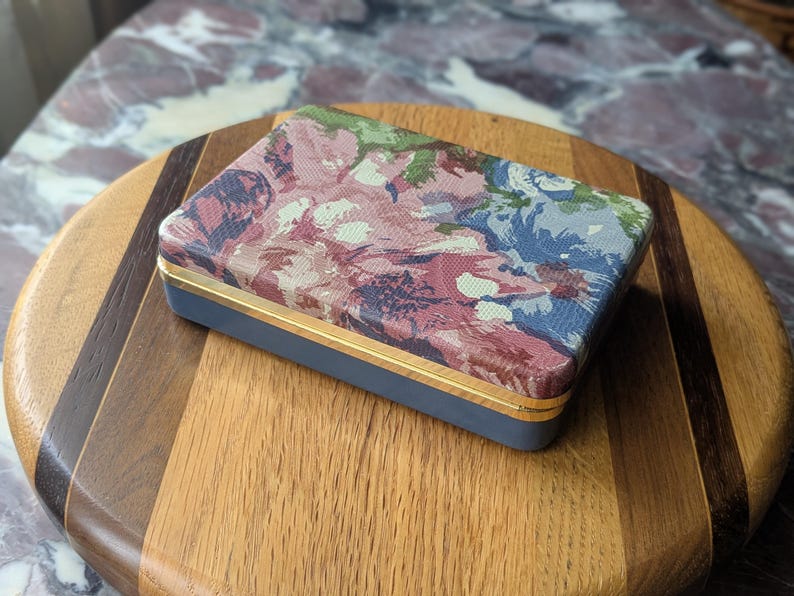 May include: A rectangular box with a floral pattern lid in shades of pink, blue, and green. The box has a navy blue base with gold trim. The box is resting on a wooden cutting board.