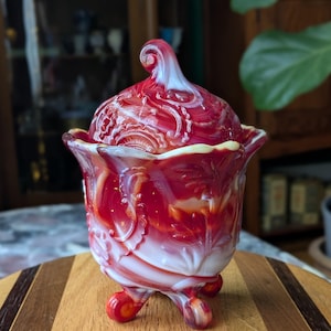 May include: A red and white glass candy dish with a lid. The dish has a ruffled edge and sits on three small feet. The lid has a swirl design and a decorative handle. The dish is approximately 6 inches tall.