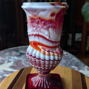 May include: A decorative glass vase with a square base and a flared top. The vase features a swirl of red, white, and orange colors. The upper portion has a relief design of figures. The vase is sitting on a wooden surface.