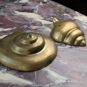 May include: Two brass decorative objects shaped like seashells. One object is a flat, circular spiral shell, and the other is a smaller, rounded, conch-style shell. Both items have a polished, metallic gold-toned finish with visible surface texture.