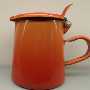 Può includere: Brocca in smalto arancione e rosso con coperchio e manico. La brocca ha un corpo arrotondato e un manico curvo. Il coperchio è incernierato e ha una piccola maniglia. La brocca è probabilmente vintage e mostra segni di usura.