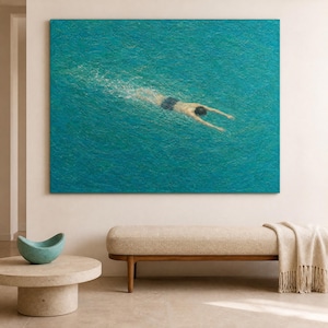 Puede incluir: Pintura al óleo de una persona nadando bajo el agua en una piscina turquesa. El nadador lleva un bañador azul oscuro. La obra de arte cuelga sobre un banco con un cojín beige y patas de madera. Una pequeña mesa redonda con un cuenco turquesa está en primer plano.