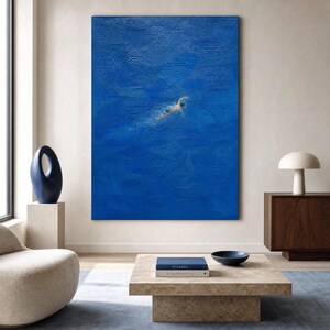 Puede incluir: Gran pintura abstracta de un nadador en una vasta extensión de agua azul. La obra de arte se exhibe en un salón moderno, con una paleta de colores neutros y una decoración minimalista. Una mesa de centro con libros y un cuenco está frente a la pintura.