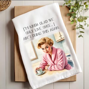 May include: White kitchen towel with the text "I'M KINDA GLAD WE ONLY LIVE ONCE. I AIN'T DOING THIS AGAIN." Features a retro illustration of a woman in a pink robe holding a coffee cup. The towel is on a wooden cutting board.