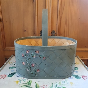 May include: A green oval-shaped basket with a woven wood base and a painted floral design. The basket has a wooden handle.