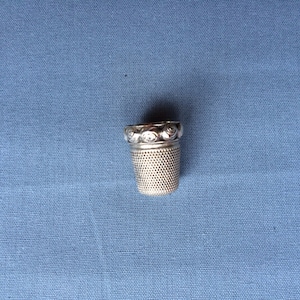 Peut inclure: Un dé à coudre en argent avec un motif de roses décoratif autour du bord supérieur. Le dé à coudre est sur un fond de tissu bleu.