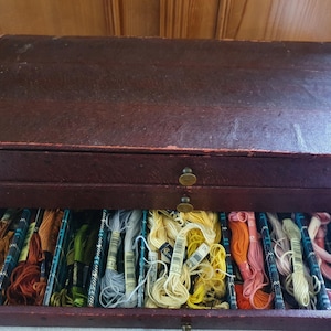 Puede incluir: Una caja de costura de madera vintage con un exterior marrón oscuro texturizado. El cajón abierto revela una colección de hilo de bordar colorido en varios tonos de amarillo, naranja, rosa, verde y azul. La caja tiene dos pequeños pomos de latón.