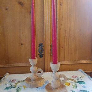May include: Two wooden candle holders with a ring handle and a red candle in each. The candle holders are on a white tablecloth with a floral pattern.