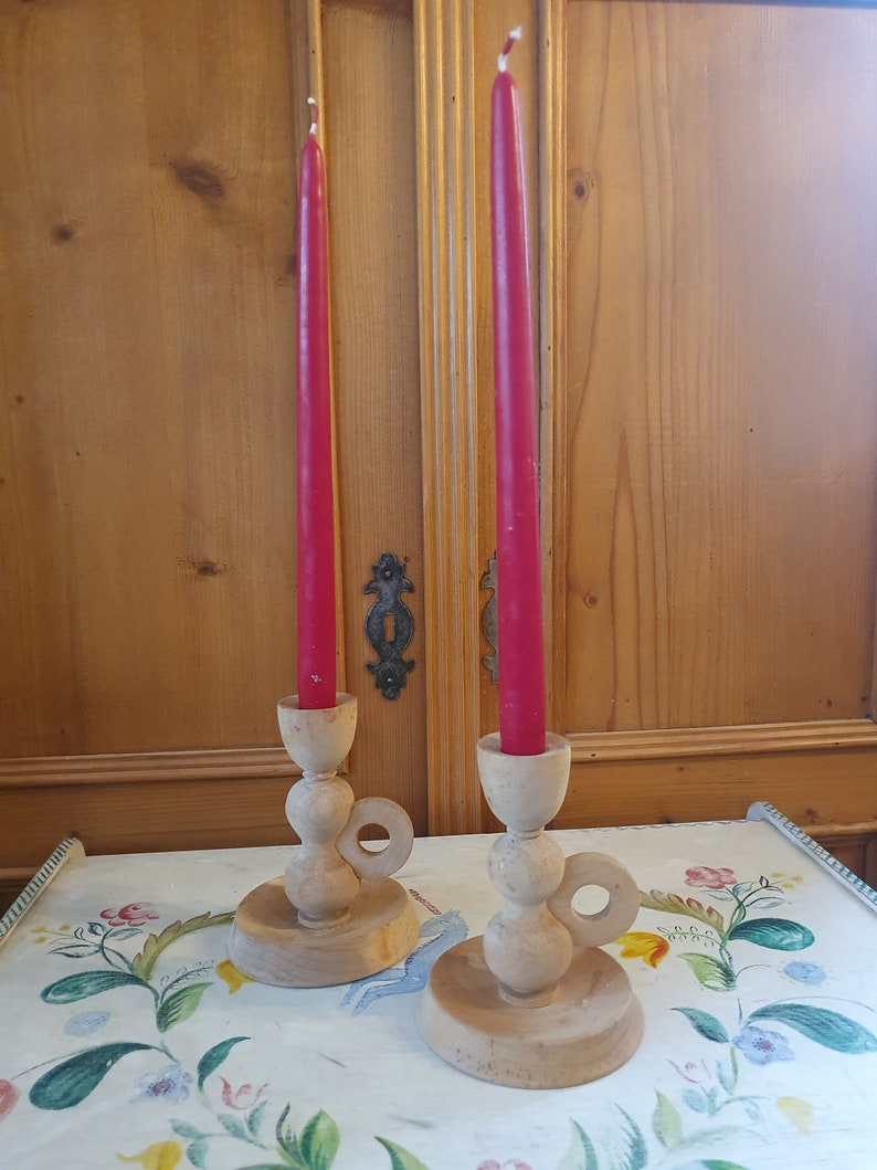 May include: Two wooden candle holders with a ring handle and a red candle in each. The candle holders are on a white tablecloth with a floral pattern.