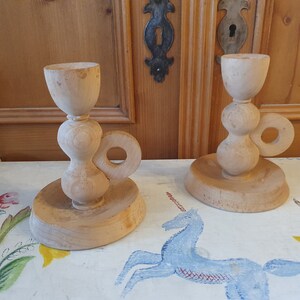 May include: Two wooden candle holders with a unique design. Each holder has a round base, a bulbous middle section, and a cup-shaped top. The holders have a ring handle on the side.