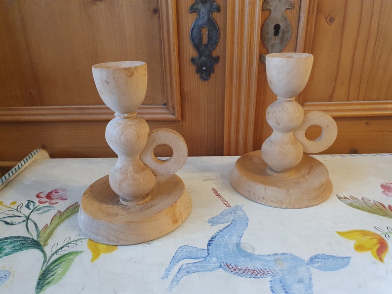 May include: Two wooden candle holders with a unique design. Each holder has a round base, a bulbous middle section, and a cup-shaped top. The holders have a ring handle on the side.