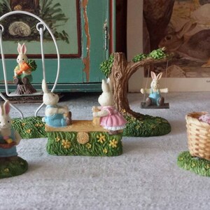 May include: A set of seven white bunny figurines with different poses and accessories. The bunnies are holding baskets, eggs, and a swing set. The figurines are on a white surface with a green background.