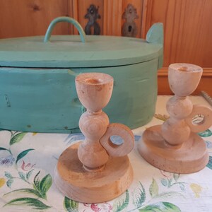May include: Two wooden candle holders with a unique design. The holders are made of natural wood and have a light brown color. The holders are on a white tablecloth with a floral pattern.
