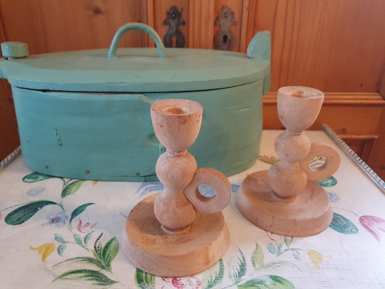 May include: Two wooden candle holders with a unique design. The holders are made of natural wood and have a light brown color. The holders are on a white tablecloth with a floral pattern.