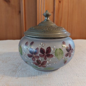 Puede incluir: Un tarro de vidrio vintage con tapa de plata, decorado con flores moradas pintadas a mano y hojas verdes sobre un fondo azul claro.