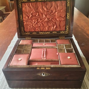 Könnte beinhalten: Ein Nähkasten aus dunklem Holz mit aufklappbarem Deckel und einer Innenauskleidung aus plissiertem, pfirsichfarbenem Satin. Das Innere enthält mehrere Fächer, einige mit pfirsichfarbenen Stoffkissen und kleinen weißen Knöpfen sowie einem Riemen.
