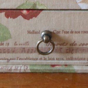 Könnte beinhalten: Eine Holzkiste im Vintage-Stil mit einem stoffbezogenen Deckel. Der Deckel zeigt ein Blumenmuster mit einer braunen und roten Rose. Die Kiste hat einen Messingring zum Öffnen mit dem Text "Meilland C'est l'une de nos roses" und "the 19 la veille. rets de sor" auf dem Deckel.