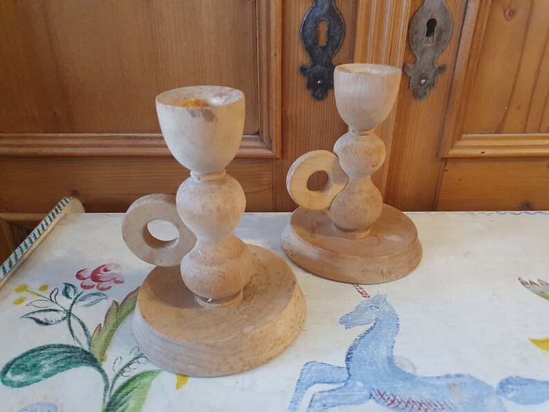 May include: Two wooden candle holders with a ring handle. The candle holders are made of light brown wood and have a simple design.
