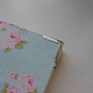 May include: A light blue fabric covered journal with a silver metal corner protector. The fabric has a floral pattern with pink roses.
