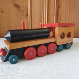 May include: A vintage wooden toy train with a black engine, red wheels, and a wooden caboose. The train has a red roof and two round windows.