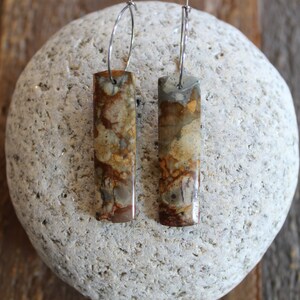 May include: A pair of silver hoop earrings with rectangular brown and beige stone pendants. The stones have a marbled pattern.