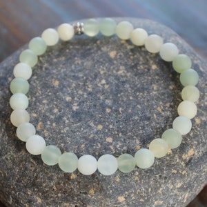 May include: A green jade stone bracelet with a silver clasp. The bracelet is made of round beads and is on a gray stone background.