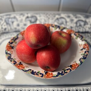 May include: Four red apples in a white bowl with a blue and gold floral pattern. The bowl is on a white surface with a black and white floral pattern.