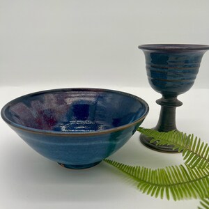 May include: A blue ceramic bowl and a goblet with a dark base. The bowl has a wide, shallow shape with a dark rim. The goblet has a fluted design and a dark, rounded base. A green fern is in the background.