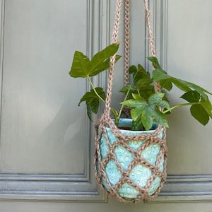 May include: A hanging plant pot with a light blue ceramic pot and a woven jute net. The pot is filled with green leafy plants. The jute rope is used to hang the pot. The background is a light green door.
