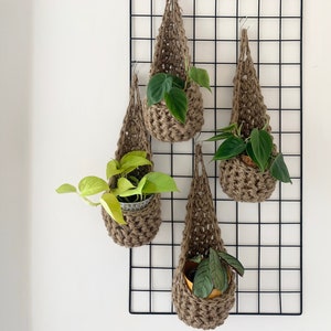 May include: Four macrame plant holders in a natural brown hue, each containing a small potted plant with green leaves. The hangers are suspended from a black grid against a white wall, creating a modern, minimalist aesthetic.