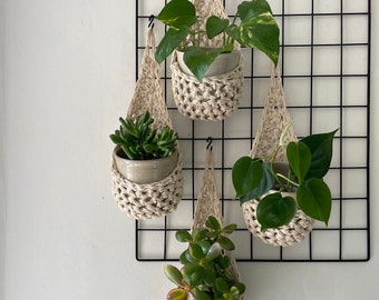 May include: Four hanging plant pots made of macrame with different plants inside. The pots are hanging from a black metal grid wall.