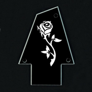 Puede incluir: Una superposici&oacute;n negra en forma de flecha para la pala de la guitarra, con un dise&ntilde;o de rosa blanca. La superposici&oacute;n tiene un borde biselado transparente y tres orificios de montaje. La rosa est&aacute; detallada con p&eacute;talos, hojas y un tallo.
