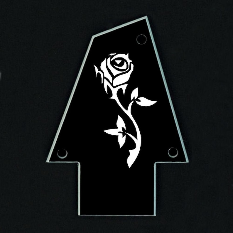 Puede incluir: Una superposici&oacute;n negra en forma de flecha para la pala de la guitarra, con un dise&ntilde;o de rosa blanca. La superposici&oacute;n tiene un borde biselado transparente y tres orificios de montaje. La rosa est&aacute; detallada con p&eacute;talos, hojas y un tallo.