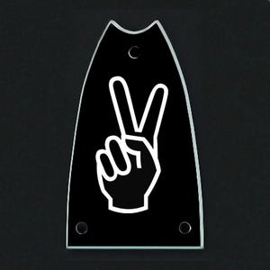 May include: A black guitar headstock plate with a white peace sign graphic. The plate has a clear, rounded top edge and three screw holes. The peace sign is a hand making the V gesture.