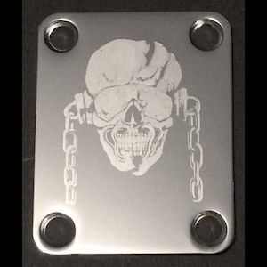 Pode incluir: Uma placa de guitarra prateada com um crânio e correntes gravados nela. O crânio tem uma expressão ameaçadora e está usando um capacete. A placa tem quatro furos para parafusos.